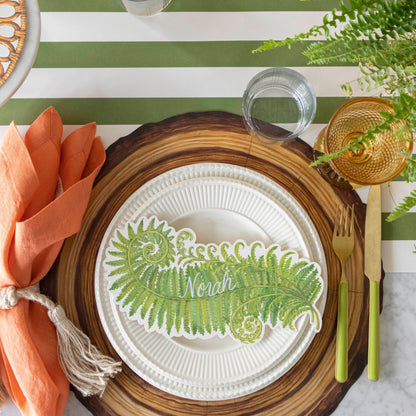 Table setting with a Die-cut Wood Slice Placemat on a green stripe runner, with white plates and a Fern table accent with the name &quot;Noah&quot; written on it on top.