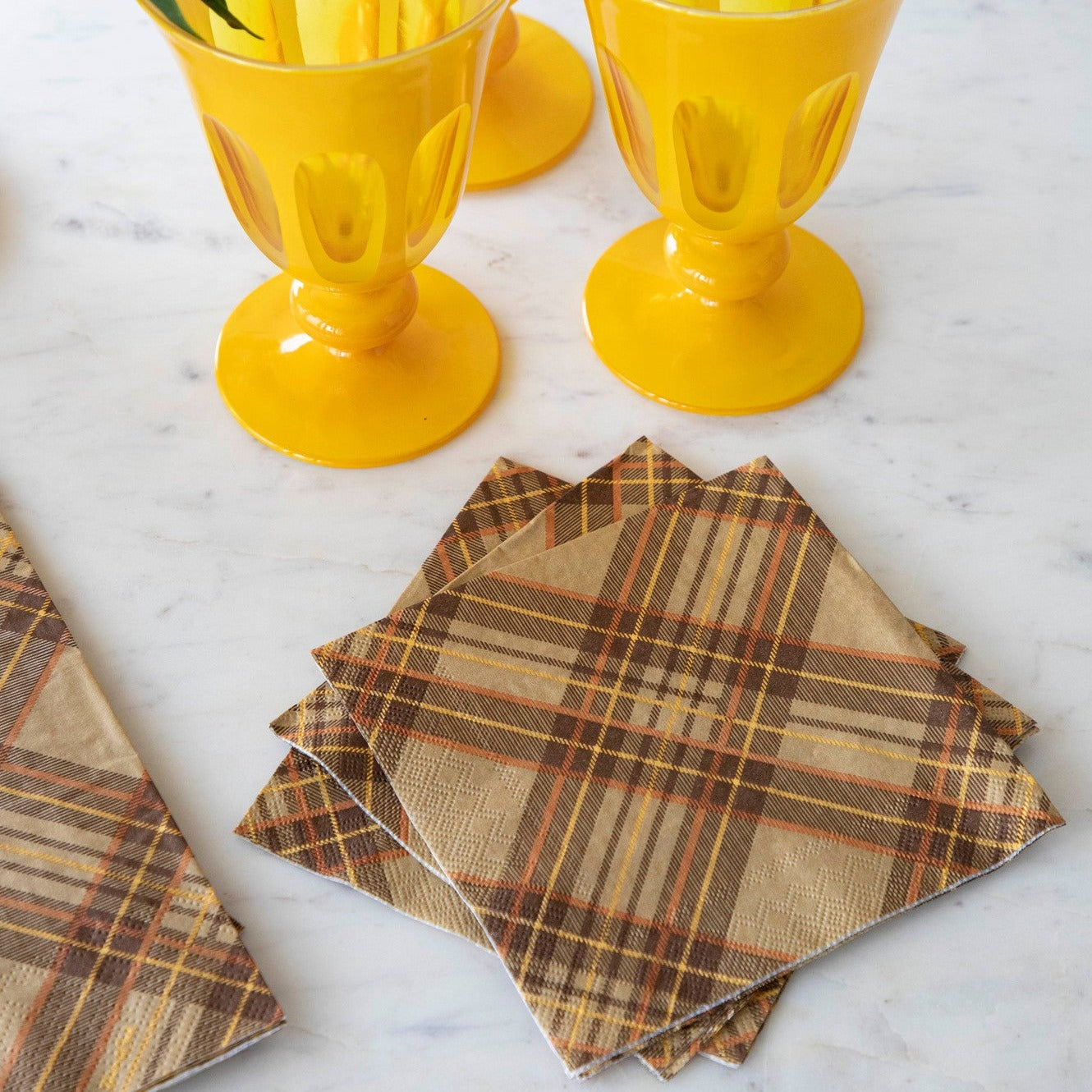 A stack of Autumn Plaid Cocktail Napkins fanned out on a white marble table with yellow glasses and an Autumn Plaid Guest Napkin.