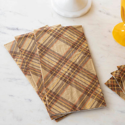 A stack of Autumn Plaid Guest Napkins fanned out on a white marble table with yellow glasses and a stack of Autumn Plaid Cocktail Napkins.