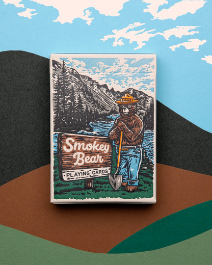 A deck of Smokey Bear Playing Cards with wildlife-themed designs, including Smokey Bear, set against a backdrop of more cards showing animal faces and fire-prevention tips. Made by Art of Play.