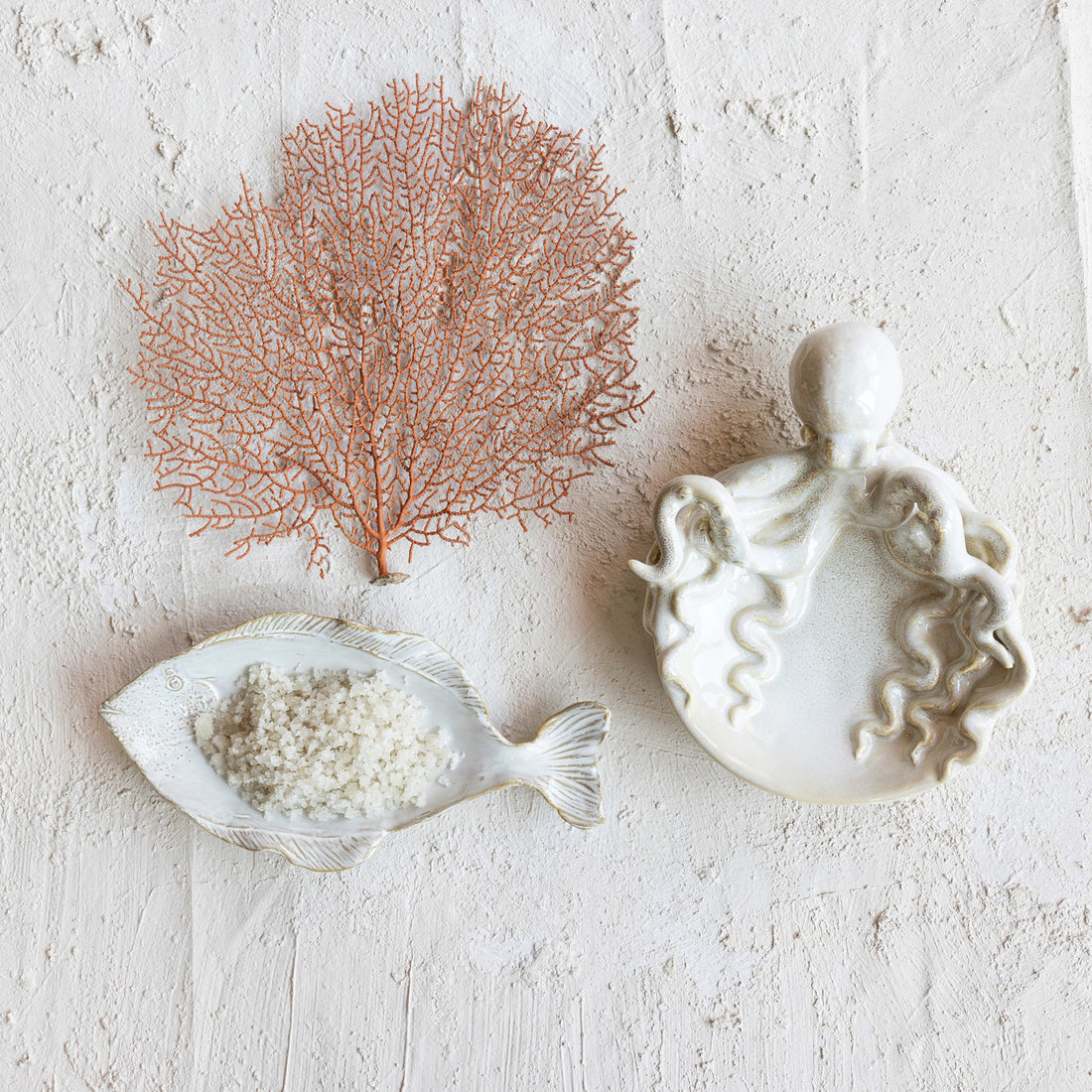 White reactive stoneware fish shaped plate on sand with an octopus shaped plate and sea algae. 