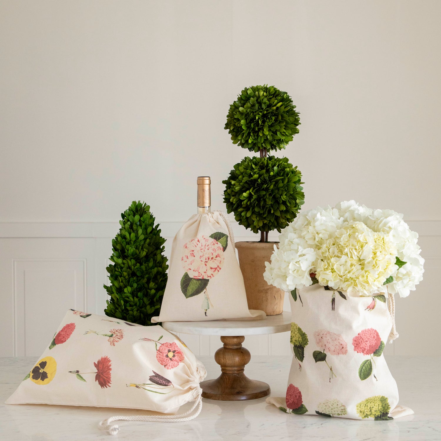 Decorative indoor setting with Laura Stoddart drawstring bags in floral patterns, topiary plants, and a vase of white hydrangeas on a table from Hester &amp; Cook.