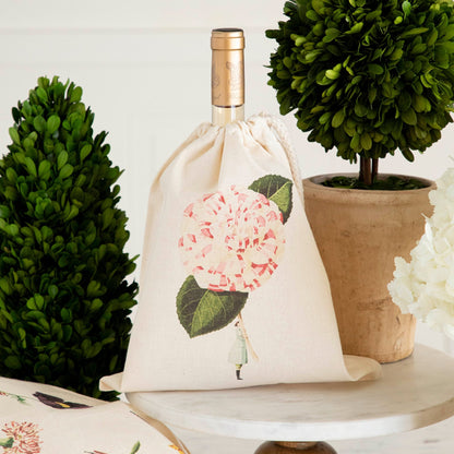 A Small Pink Peony Laura Stoddart Drawstring Bag containing a bottle of wine, sitting on a pedestal with potted plants. 