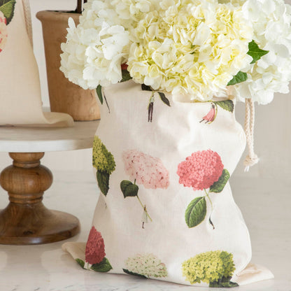 A Medium Hydrangeas Laura Stoddart Drawstring Bag sitting on a white table containing white flowers.