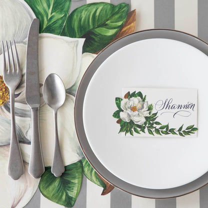 An elegant table setting with the Magnolia Place Card with &quot;Shannon&quot; written on it, on a set of dinner plates, next to the Die-cut Magnolia Placemat and Silver Classic Stripe Runner underneath.