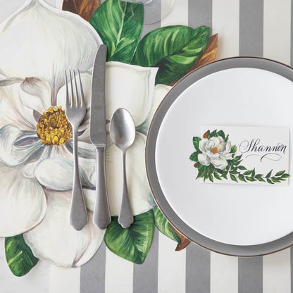 Die-cut Magnolia Placemat with silverware on top, next to a table setting with a Magnolia Place Card with the name &quot;Shannon&quot; written on it.