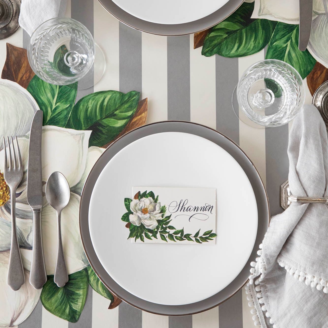 An elegant table setting with the Magnolia Place Card with &quot;Shannon&quot; written on it, on a set of dinner plates, next to the Die-cut Magnolia Placemat and Silver Classic Stripe Runner underneath.