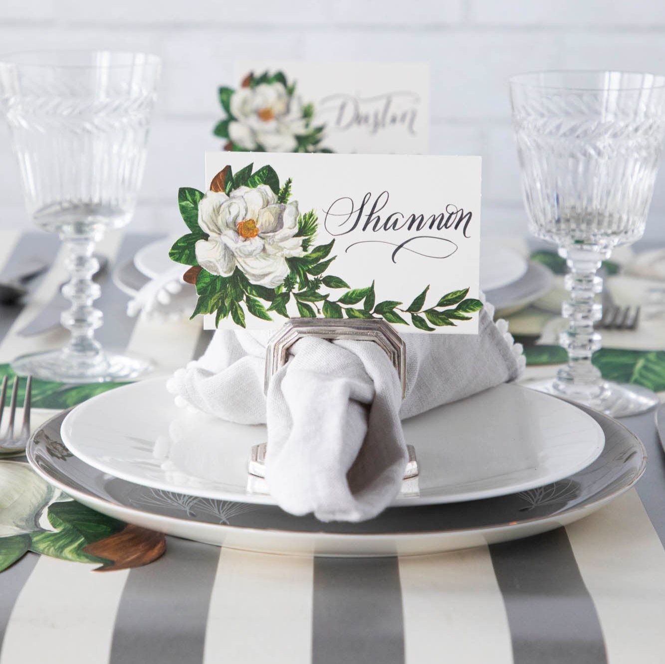 An elegant table setting with the Magnolia Place Card with &quot;Shannon&quot; written on it, inside of a silver napkin ring place card holder.