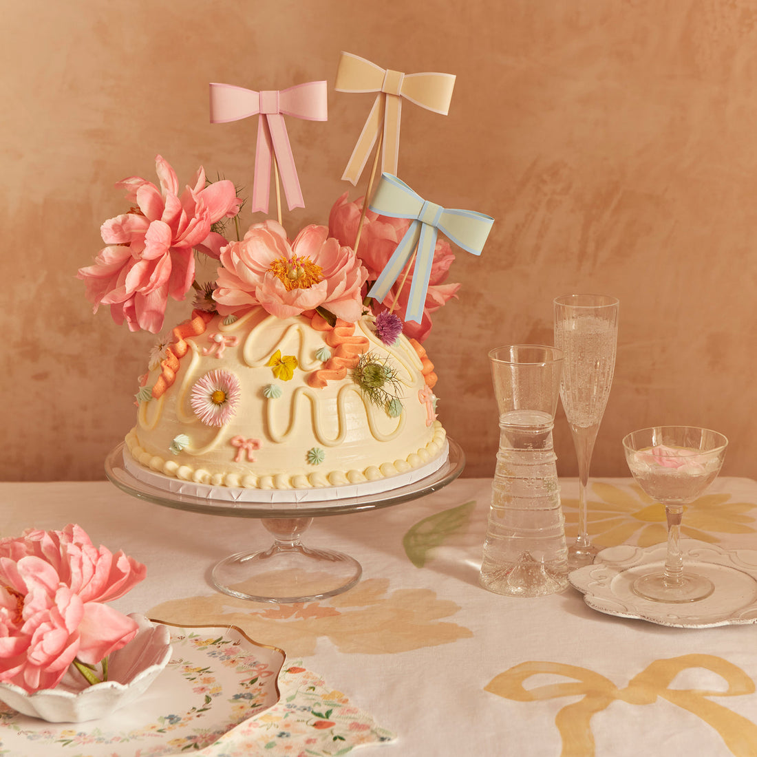 Bow cake toppers shown on a cake adorned with flowers. 