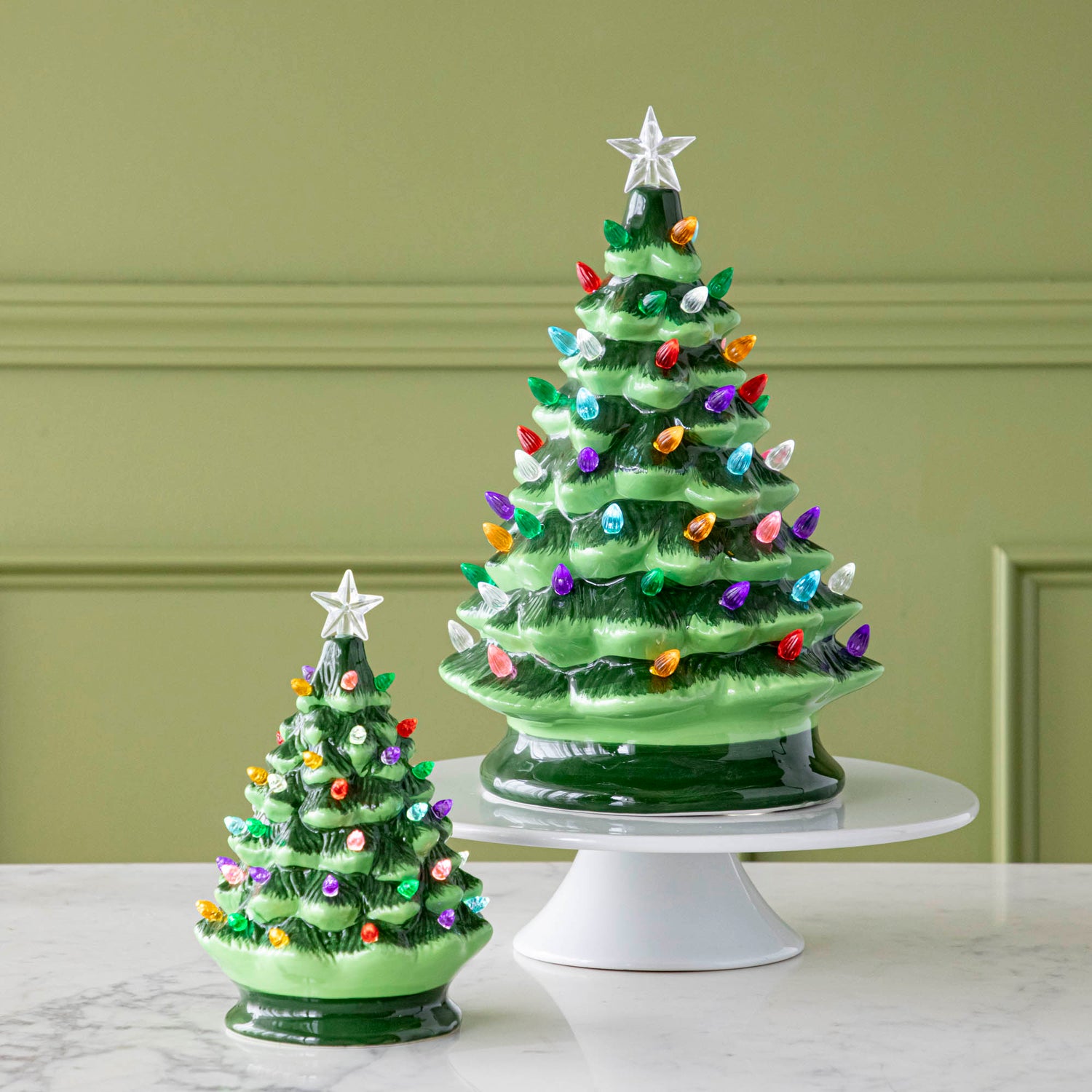 Two Porcelain Vintage Lit Trees with Timers; one on a cake stand and one on a marble table.