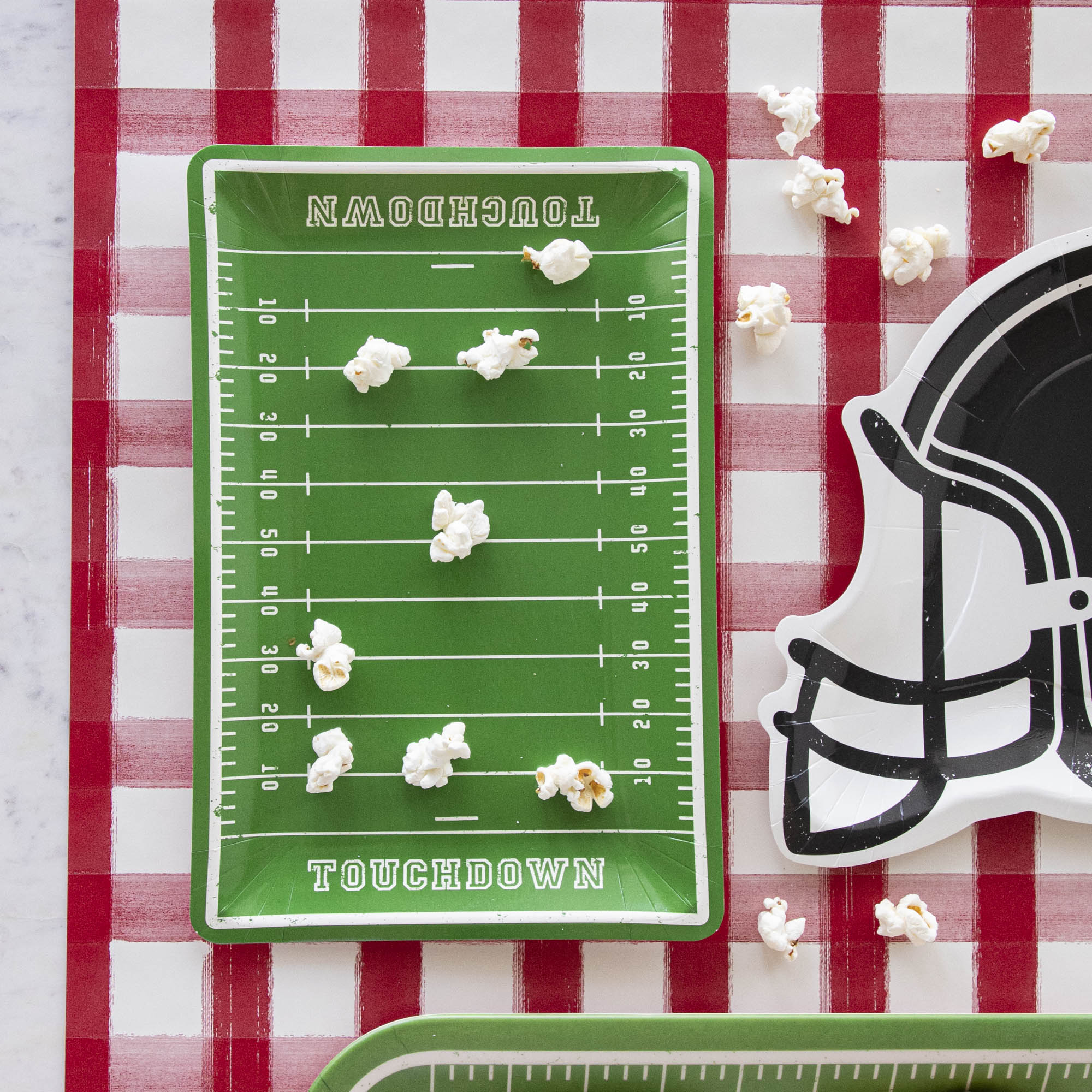 A football field with Football Field Paper Plates and a football helmet on a red and white checkered tablecloth, accompanied by game day goodies.