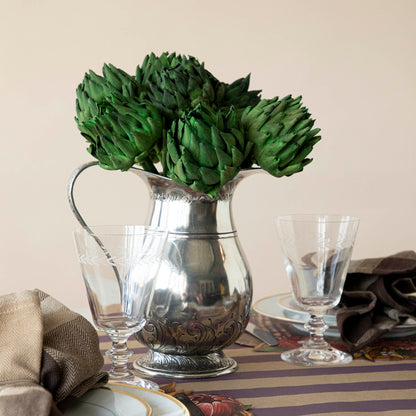 Dried Green Artichokes shown in silver vase on a table with glasses and place settings. 