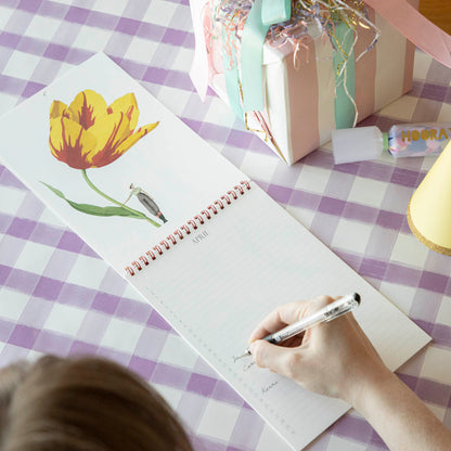 The Laura Stoddart Birthday Calendar resting on a table, opened up to the April page, with a woman writing names on the blank lines next to the dates.