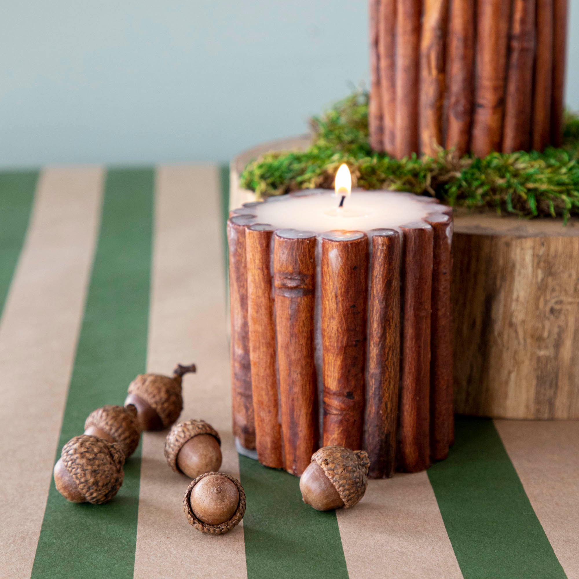 A Cinnamon Stick Scented Pillar Candles lit up, next to acorns and a wooden stump.