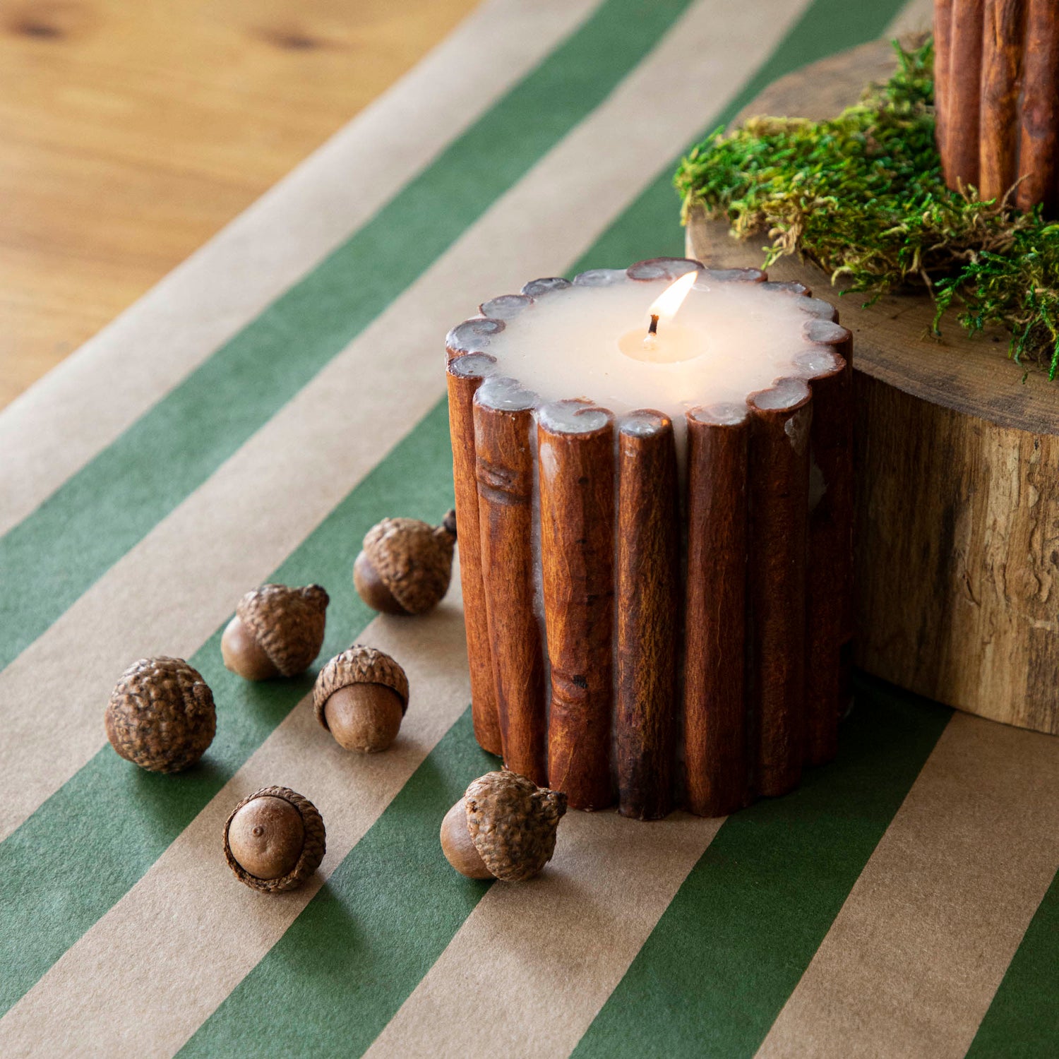 The lit Cinnamon Stick Scented Pillar Candle next to acorns, on top of the Kraft Green Classic Stripe Runner.