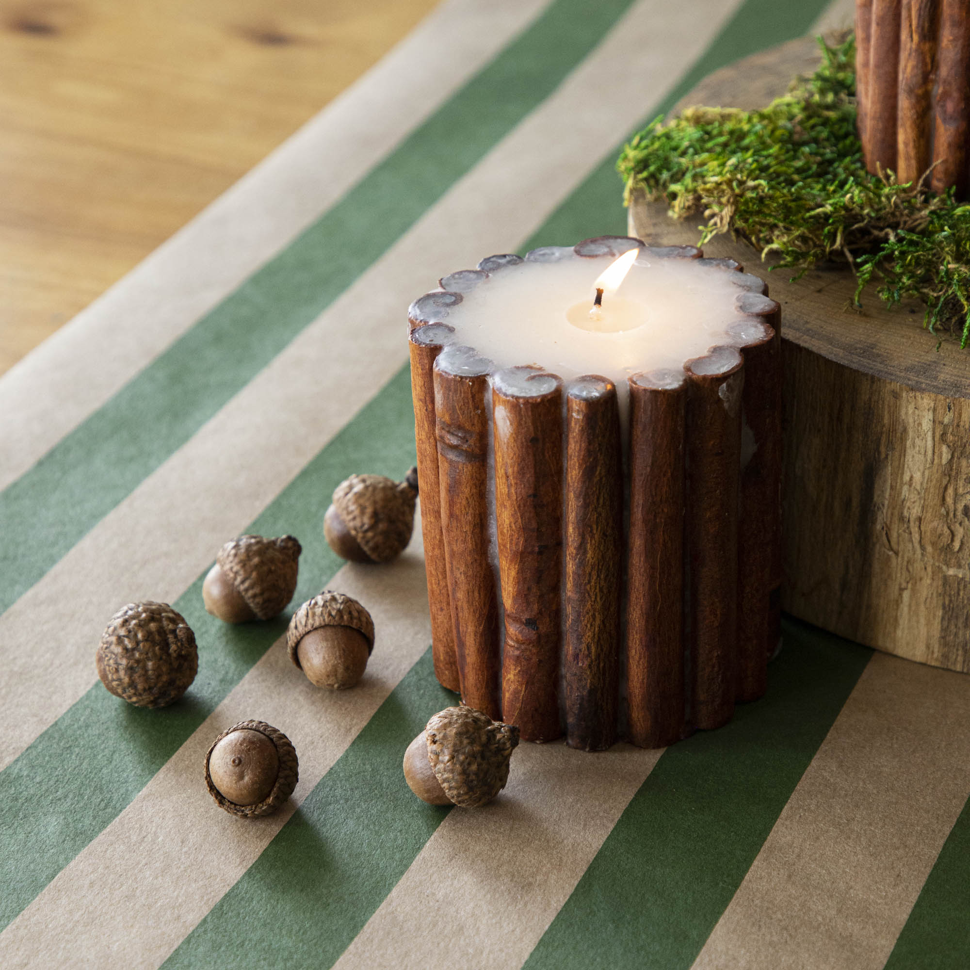 The lit Cinnamon Stick Scented Pillar Candle next to acorns, on top of the Kraft Green Classic Stripe Runner.