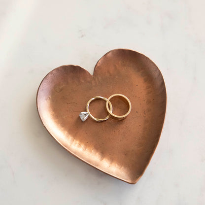 A HomArt Forged Iron Heart Tray with gold and silver rings on a copper-colored, pink, and white striped surface.