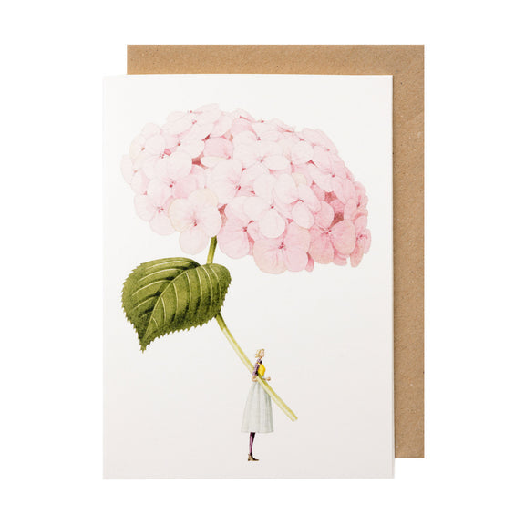 A white greeting card featuring a stylized illustration of a woman holding a gigantic light pink hydrangea bloom by the stem, with the included kraft paper envelope tucked behind. 