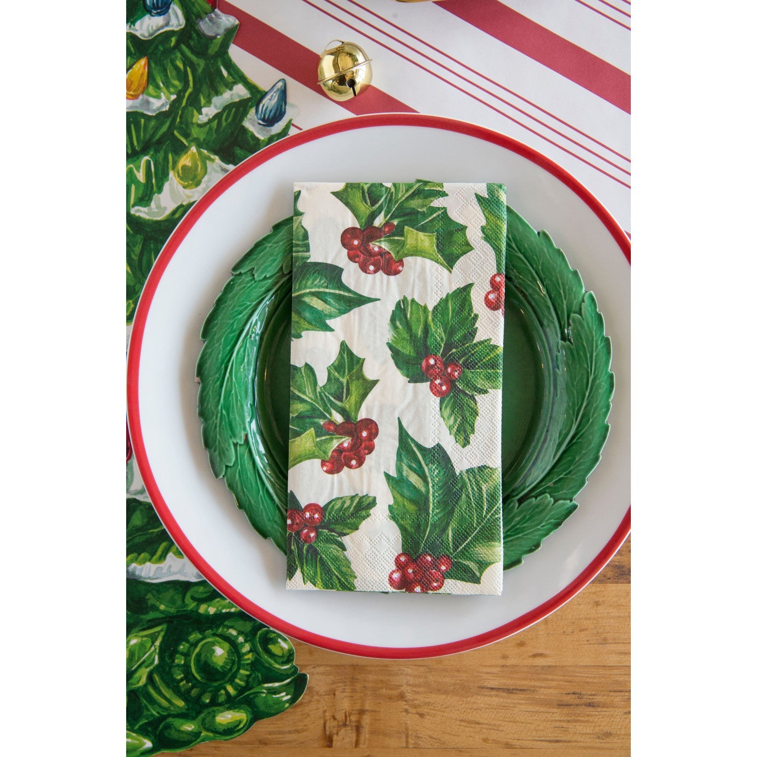 A square cocktail napkin featuring painterly green holly leaves and red berries in the lower right, with decorative gold frame lines along the lower and right edges, on a white background.