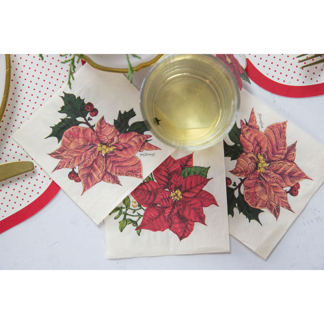 Three Poinsettia Cocktail Napkins spread on a table, showing the red front design and the pink back design, next to a glass of white wine.