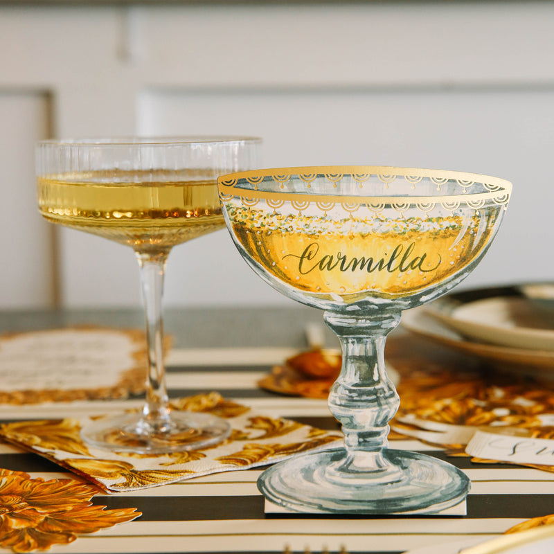 An elegant table setting with a Champagne Coupe Place Card reading &quot;Carmilla&quot; standing next to a real coupe of champagne.