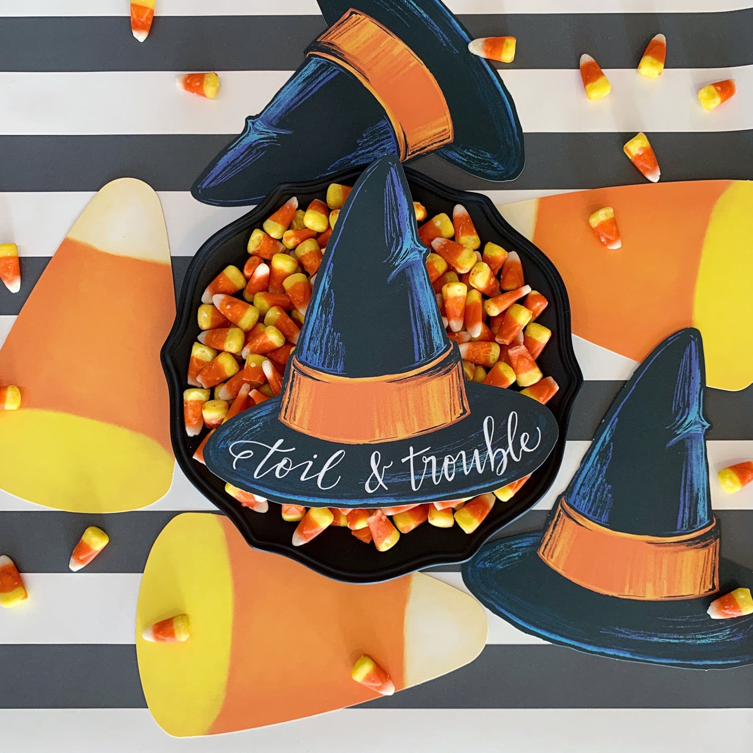 Top-down view of a Witch Hat Table Accent with &quot;toil &amp; trouble&quot; written on it resting on a big bowl of candy corn, on a treat table surrounded by other Halloween table accents.