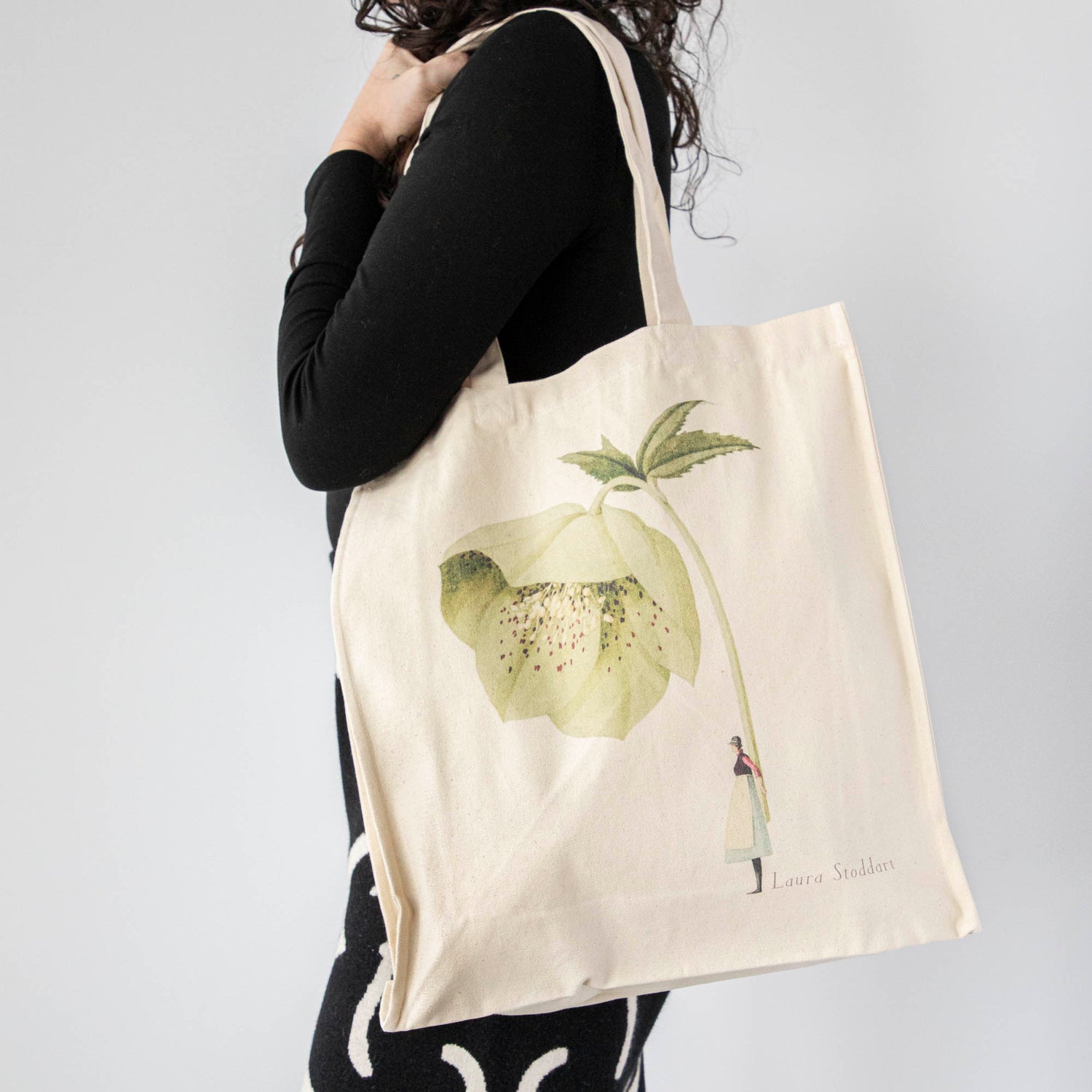 A close-up of the Hellebore Heavyweight Canvas Tote Bag being carried over the shoulder of a woman in black, showing the large functional size of the bag.
