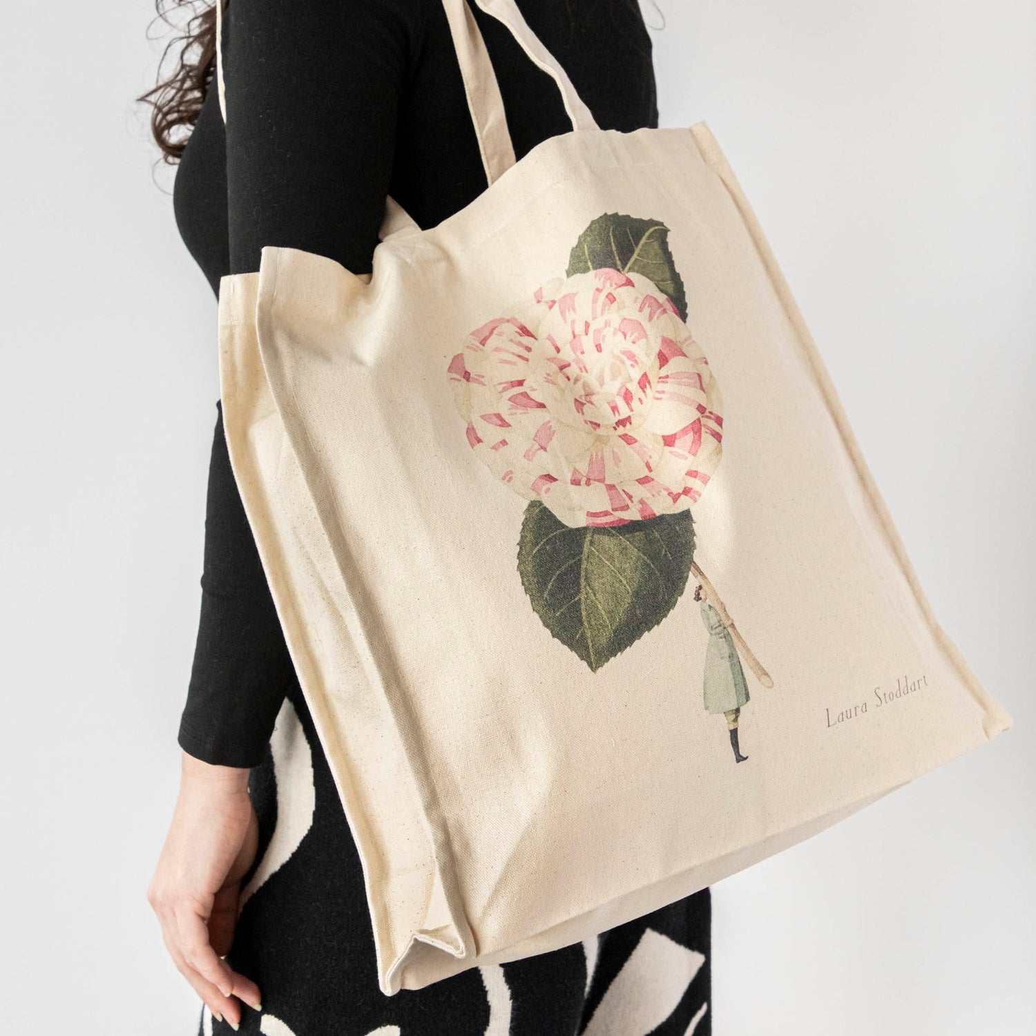 A close-up of the Camelia Heavyweight Canvas Tote Bag being carried over the shoulder of a woman in black, showing the large functional size of the bag.