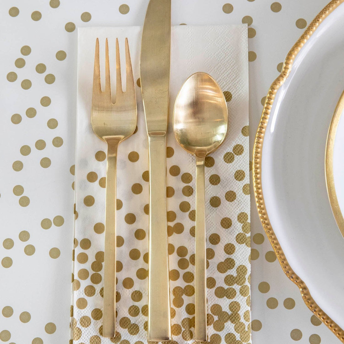 A Gold Confetti Guest napkin under gold cutlery next to a gold-rimmed plate, on a gold confetti table runner.