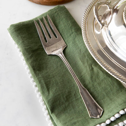 A Hester &amp; Cook vintage silver-plate meat fork resting on a folded green napkin with pom-pom trim on a marble countertop.