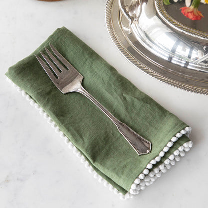 A Hester &amp; Cook vintage silver-plate meat fork resting on a folded green napkin with pom-pom trim on a marble countertop.