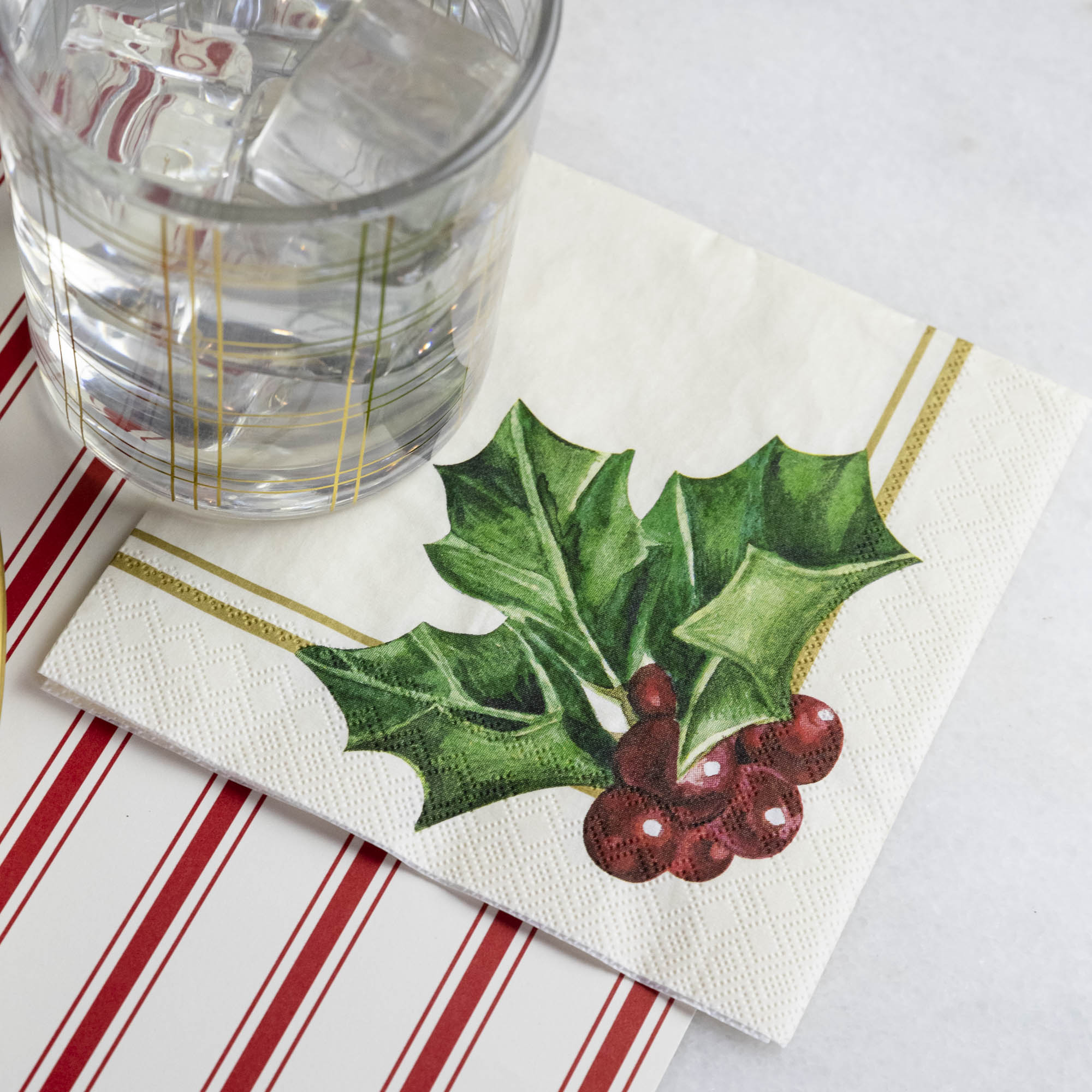 A square cocktail napkin featuring painterly green holly leaves and red berries in the lower right, with decorative gold frame lines along the lower and right edges, on a white background.