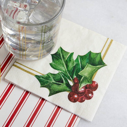 A square cocktail napkin featuring painterly green holly leaves and red berries in the lower right, with decorative gold frame lines along the lower and right edges, on a white background.