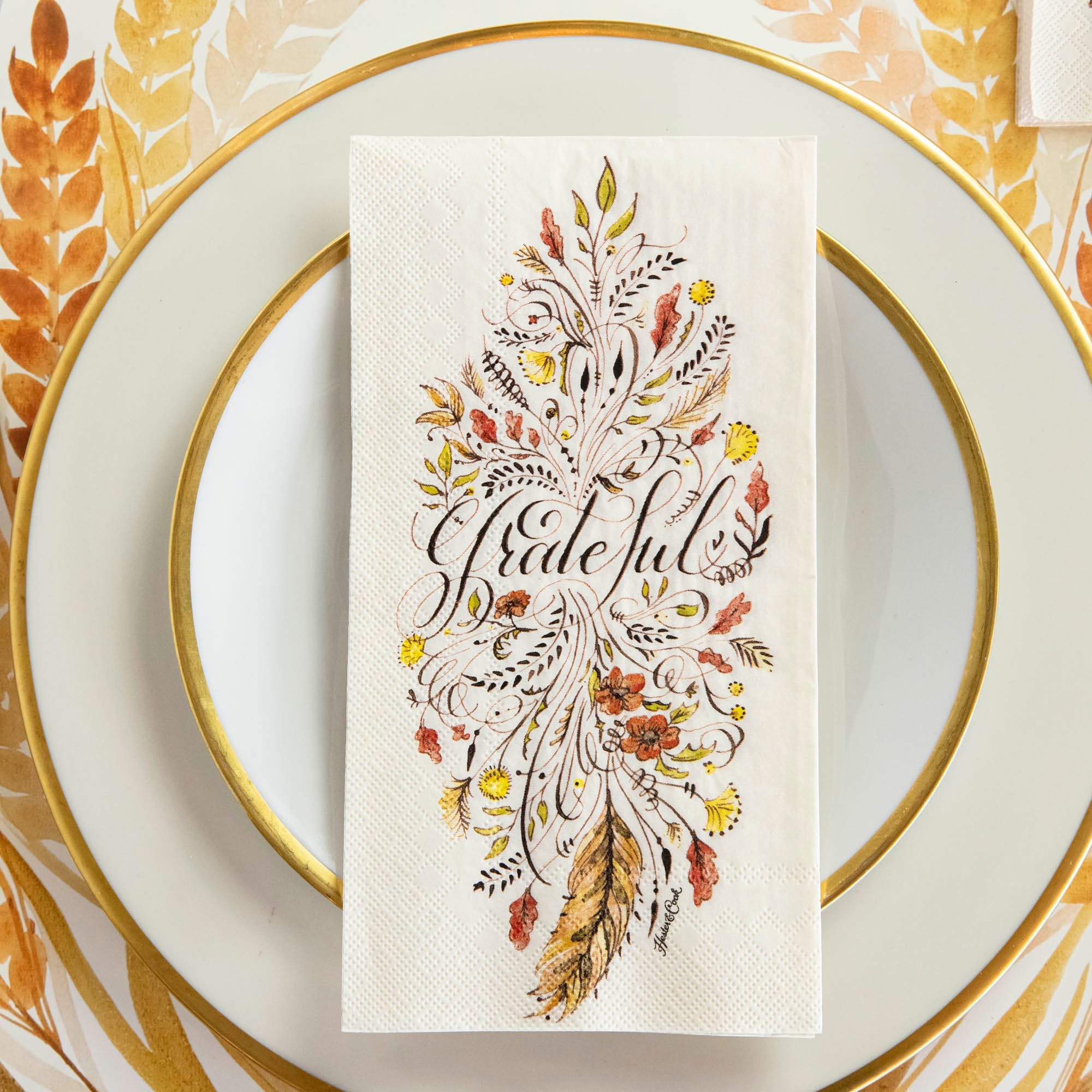 Top-down view of an elegant Thanksgiving place setting, featuring a Grateful Guest Napkin centered on a gold-rimmed plate.