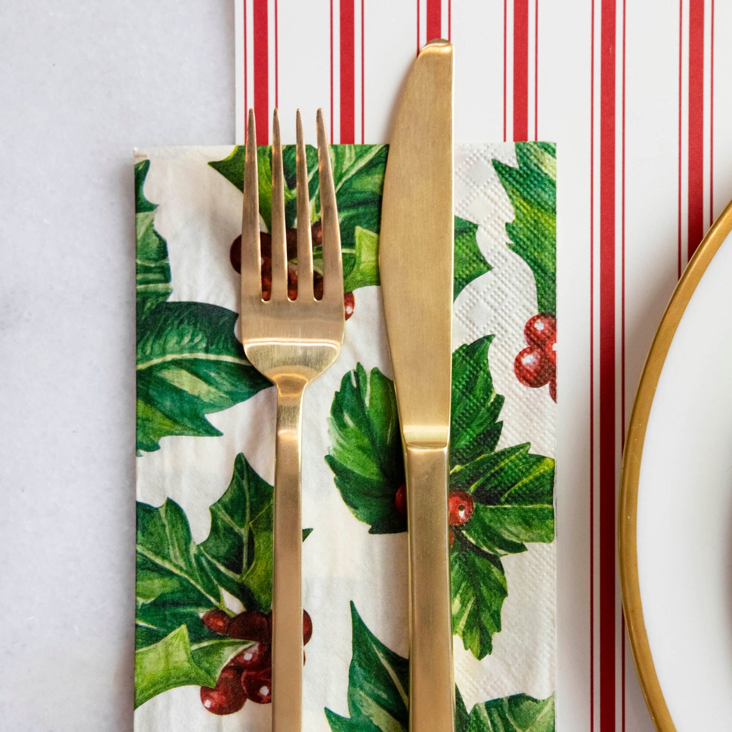 A square cocktail napkin featuring painterly green holly leaves and red berries in the lower right, with decorative gold frame lines along the lower and right edges, on a white background.