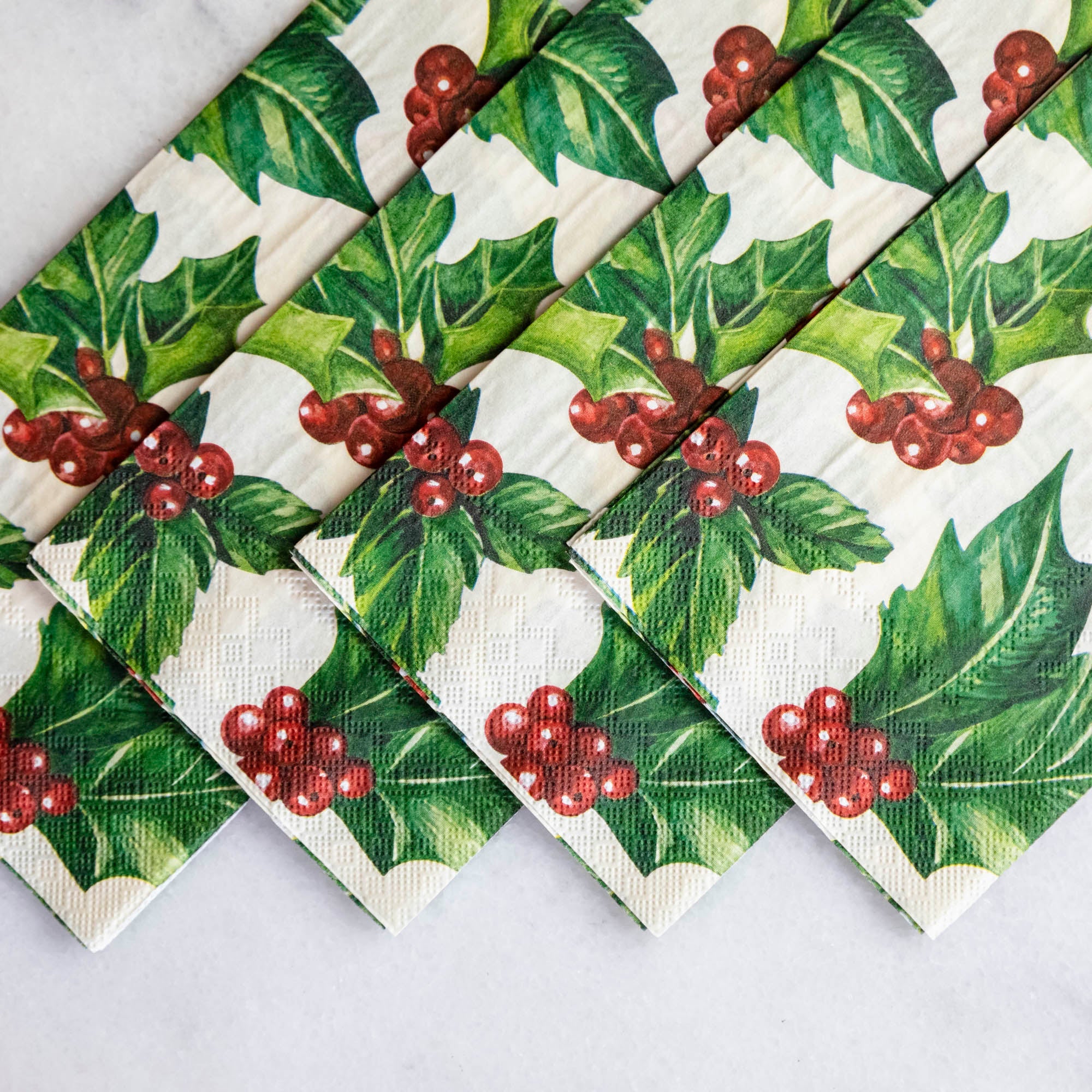 A square cocktail napkin featuring painterly green holly leaves and red berries in the lower right, with decorative gold frame lines along the lower and right edges, on a white background.