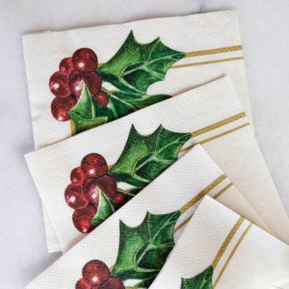 A square cocktail napkin featuring painterly green holly leaves and red berries in the lower right, with decorative gold frame lines along the lower and right edges, on a white background.