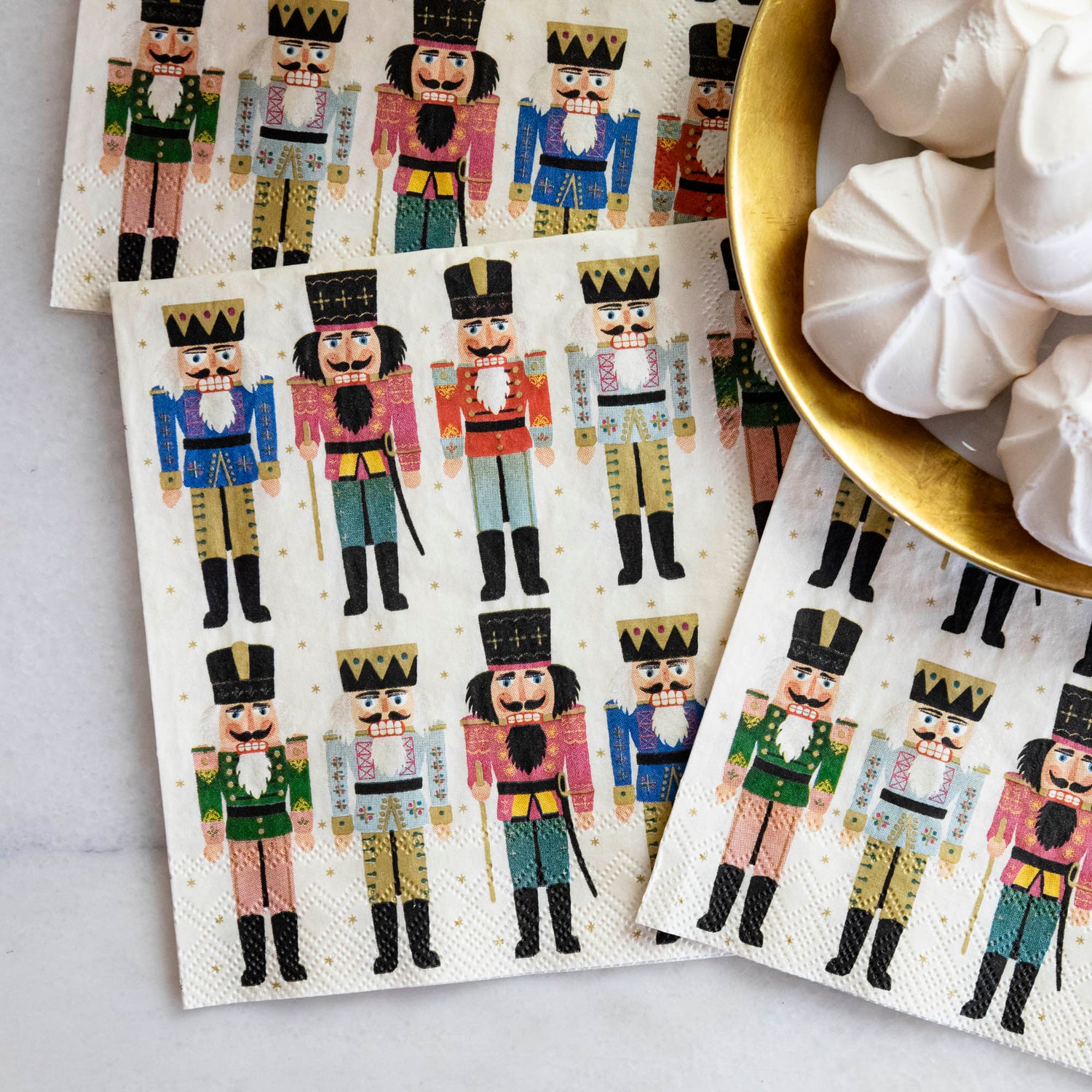 Close-up of three Nutcracker Cocktail Napkins tucked under a gold-rimmed plate of treats.