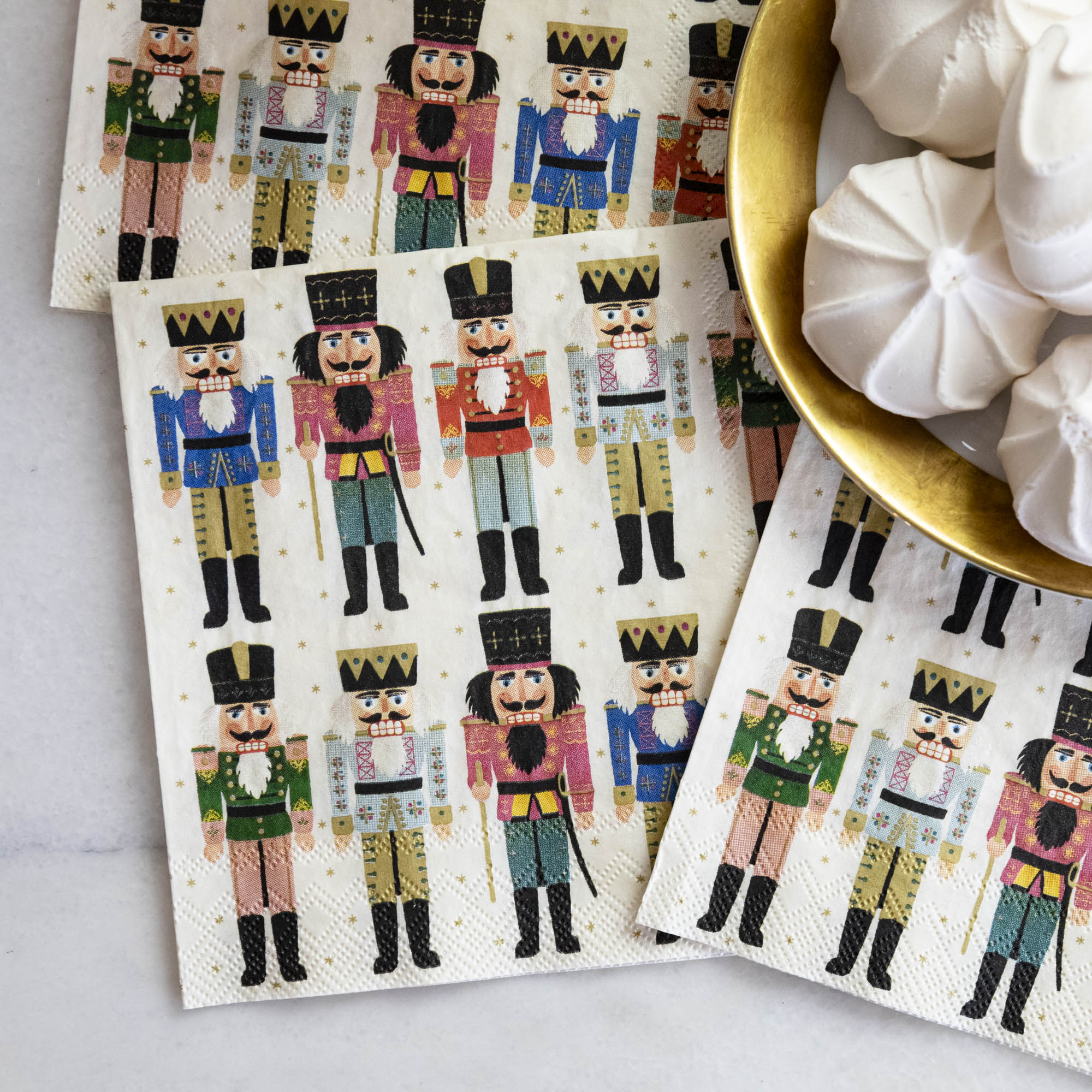 Close-up of three Nutcracker Cocktail Napkins tucked under a gold-rimmed plate of treats.