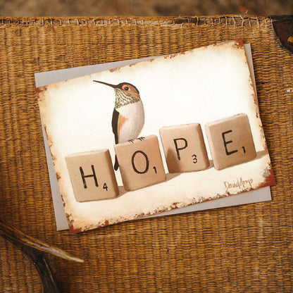 A &quot;Hope&quot; (Scrabble) notecard with a hummingbird illustration and the word &quot;hope&quot; spelled out with letter tiles rests on a woven surface. (David Arms)