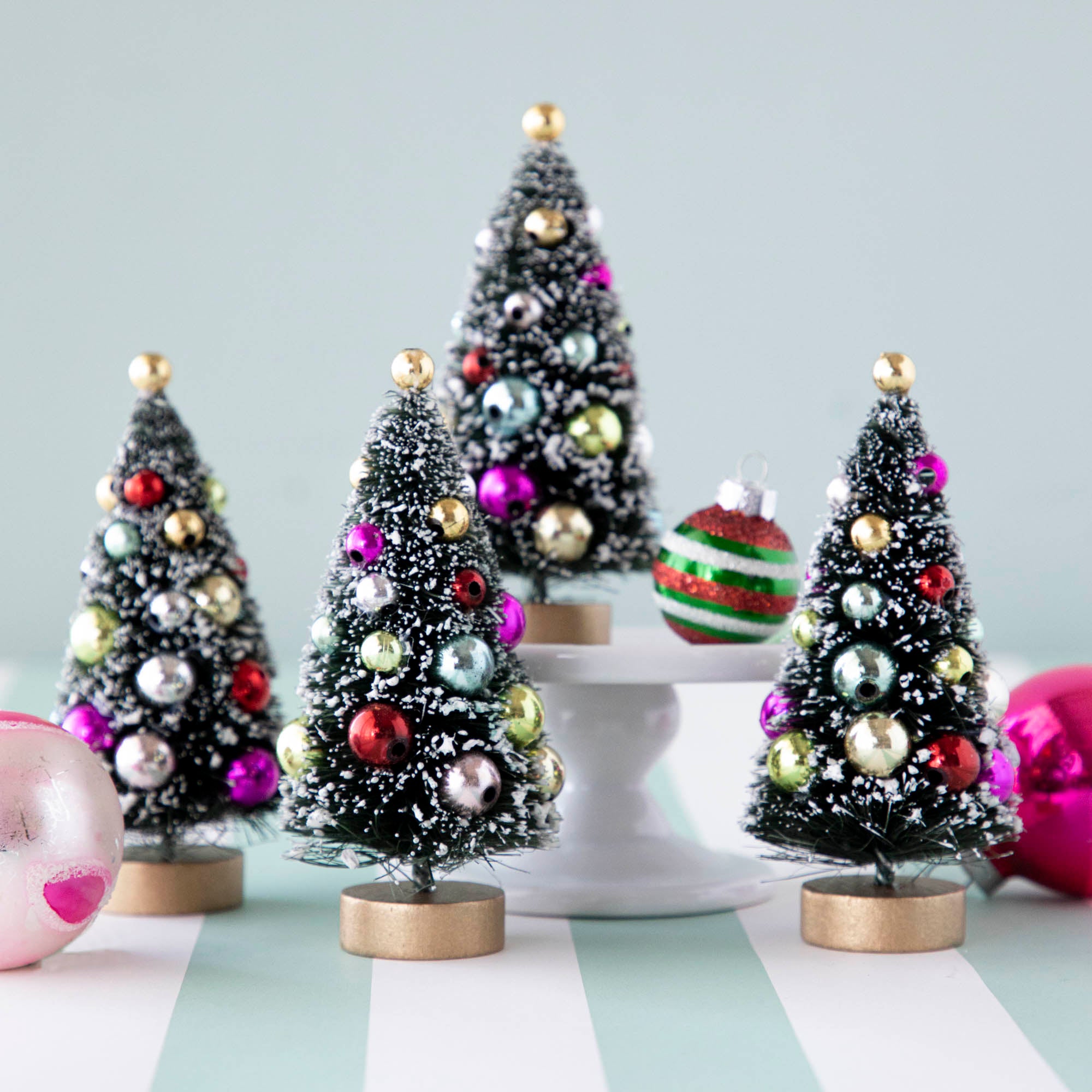 Four, 4&quot; holiday sisal trees with colorful ornaments and snow, displayed on a table with ornaments.