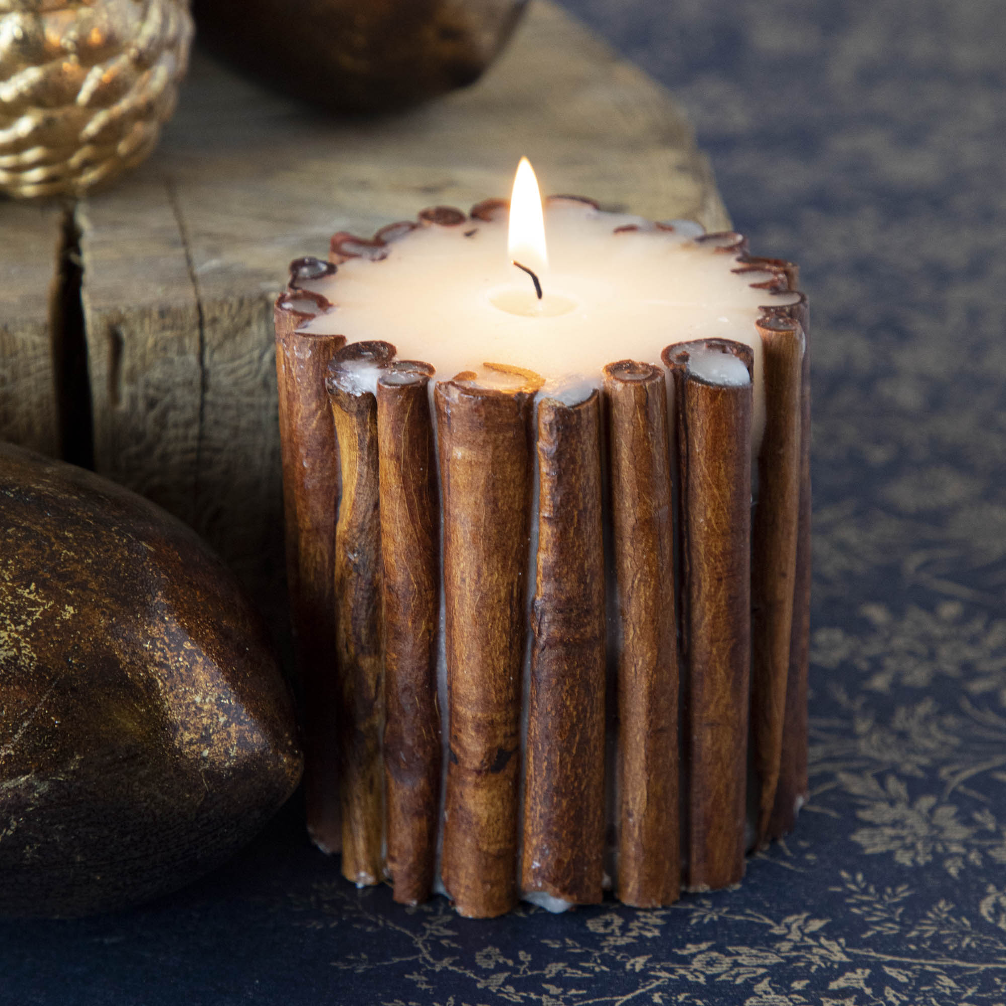 The small Cinnamon Stick Scented Pillar Candle lit, propped next to brass acorns and a tree stump.